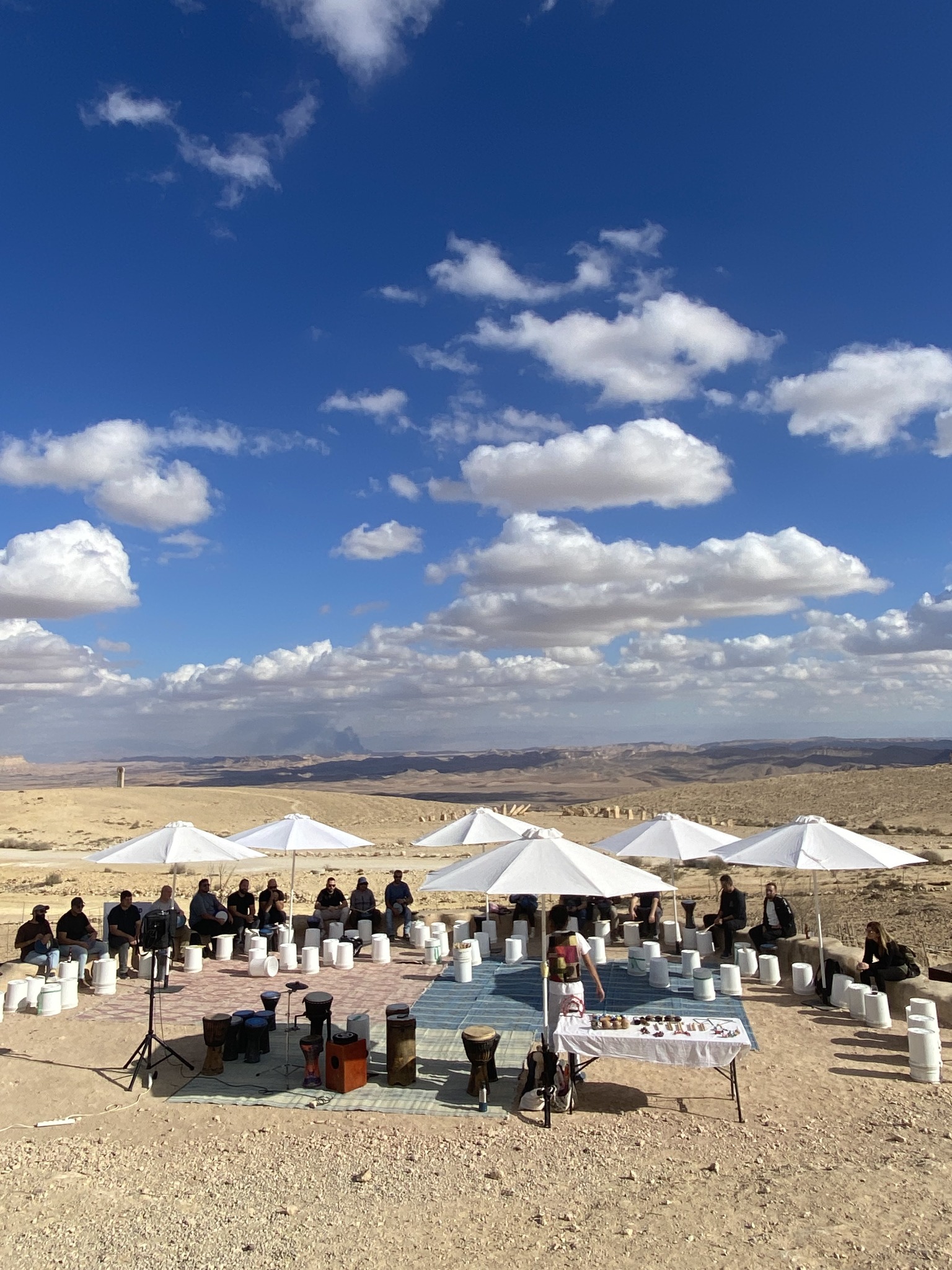 משתתפים יושבים במעגל עם כלי הקשה תחת שמשיות במדבר.
