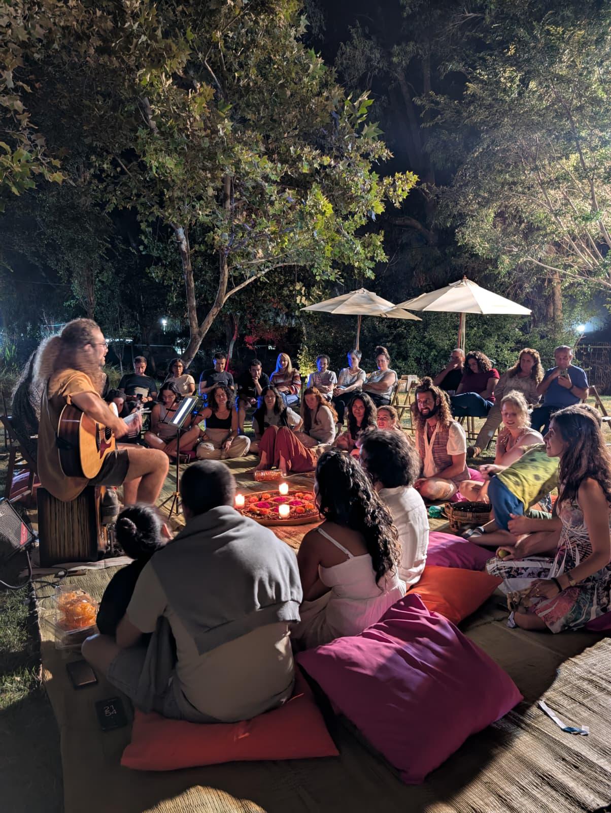 מפגש שירה משותפת בחוץ, עם גיטרה אקוסטית ומשתתפים שיושבים במעגל.